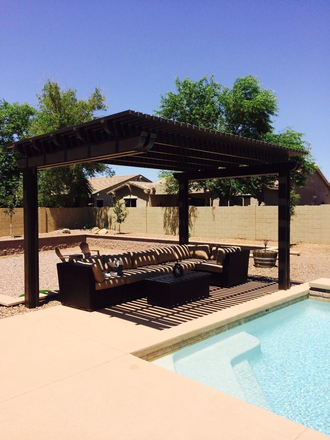 Pergola with seating area near a pool. Brown wood structure casts shadows; clear blue sky.