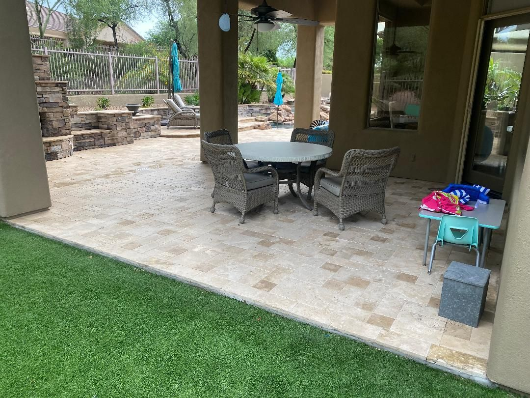 Patio with stone pavers, table, chairs, and artificial turf lawn. Beige and green colors.