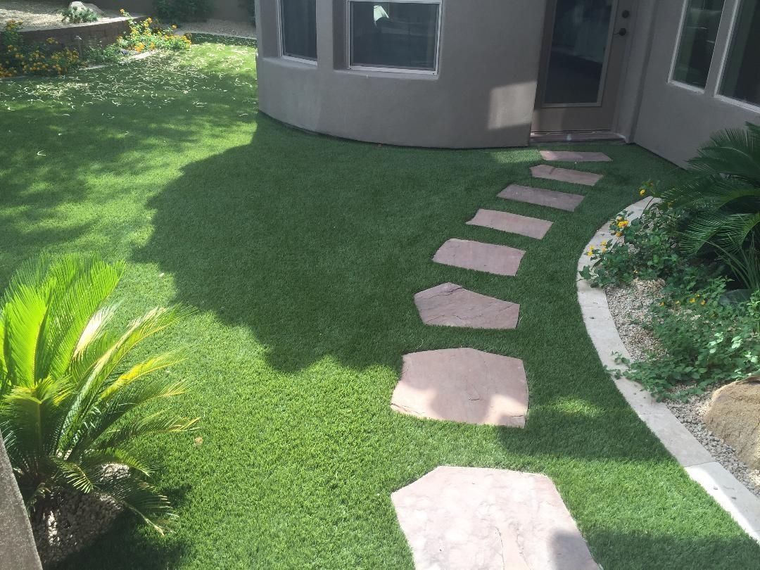 Stone path through artificial grass leading to a house entrance. Palm-like plants on the left, building on the right.