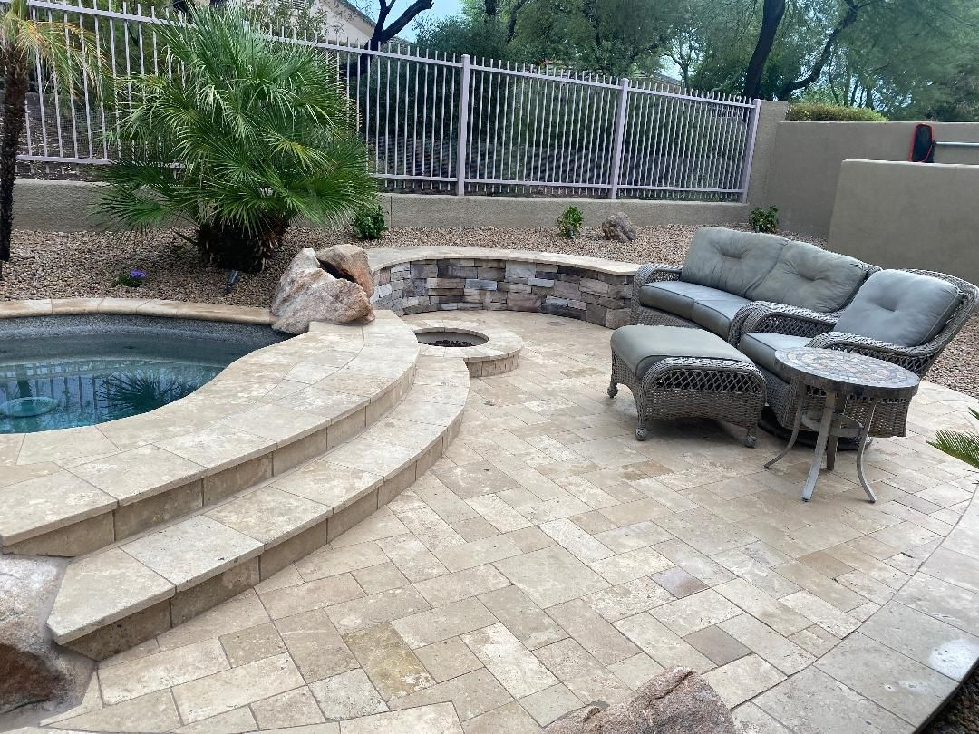 Patio with pool, seating area, fire pit, stone pavers. Gray wicker furniture, tan stone accents, lush greenery.