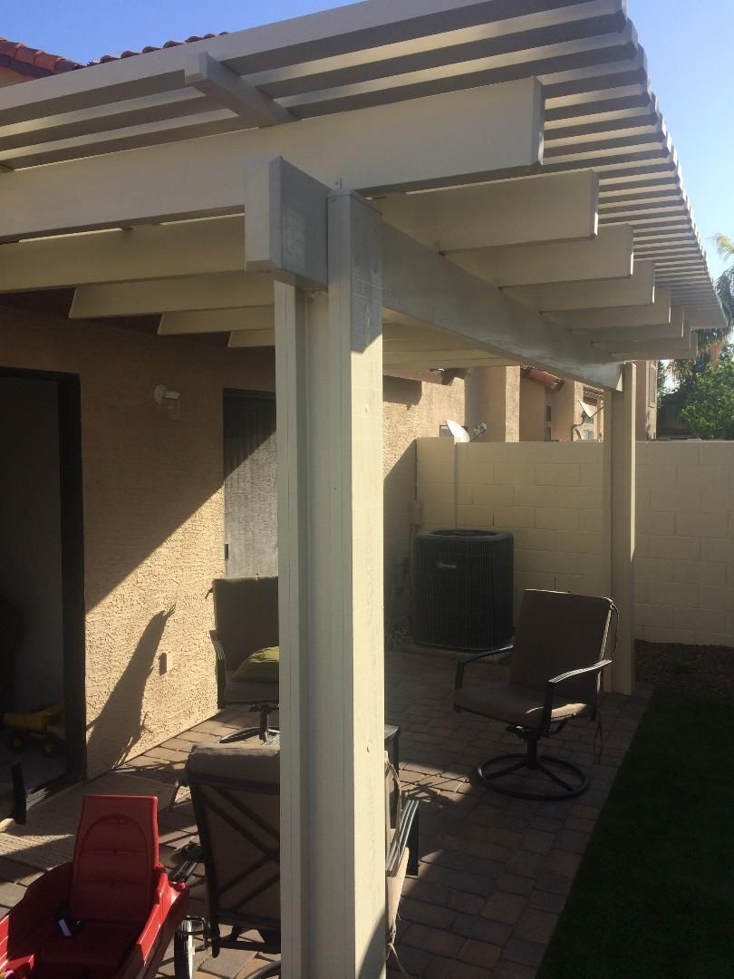 Beige pergola over a patio with chairs and a wall. Sunlight casts shadows.