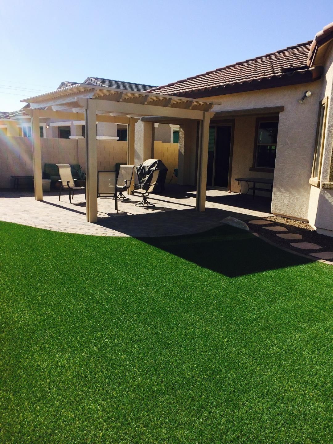 Patio with a wooden pergola, chairs, grill, and artificial green grass under a blue sky.