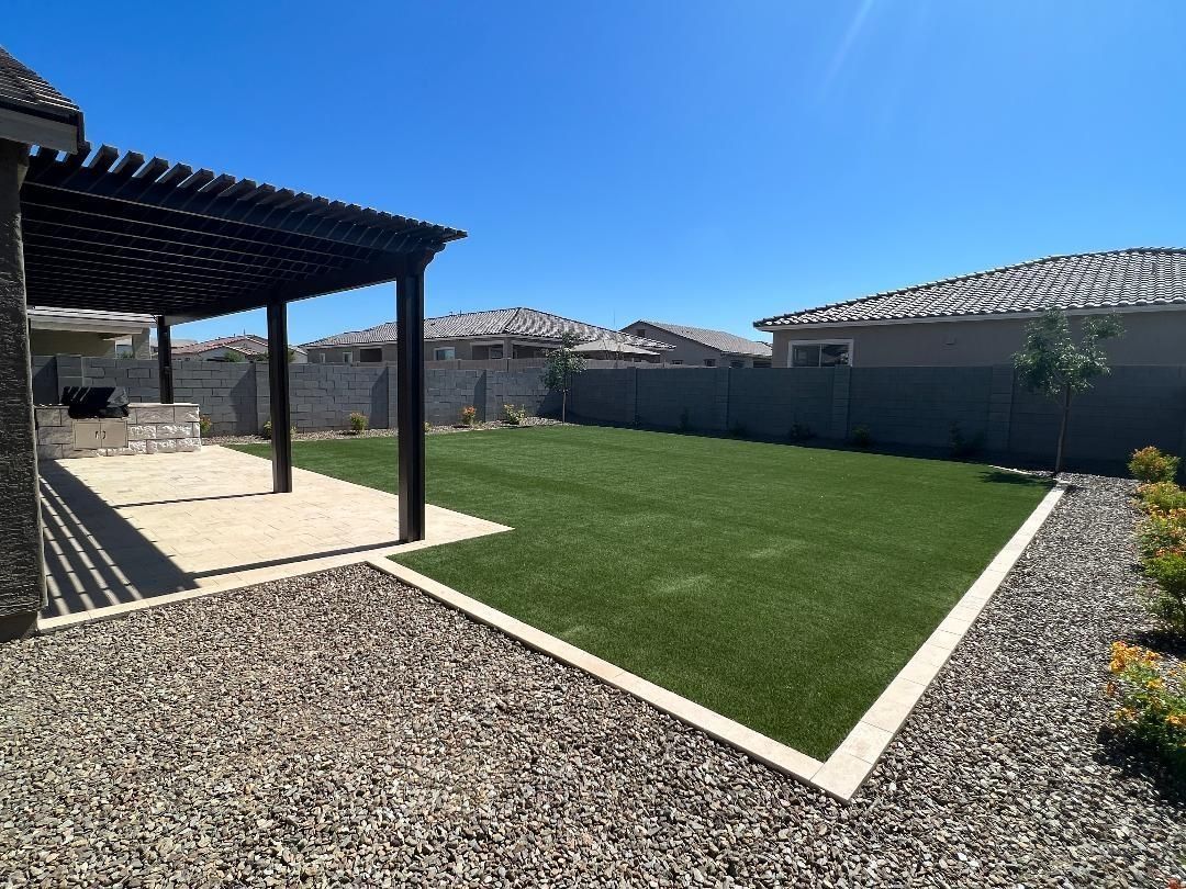 Backyard with pergola, artificial grass lawn, gravel border, and blue sky.