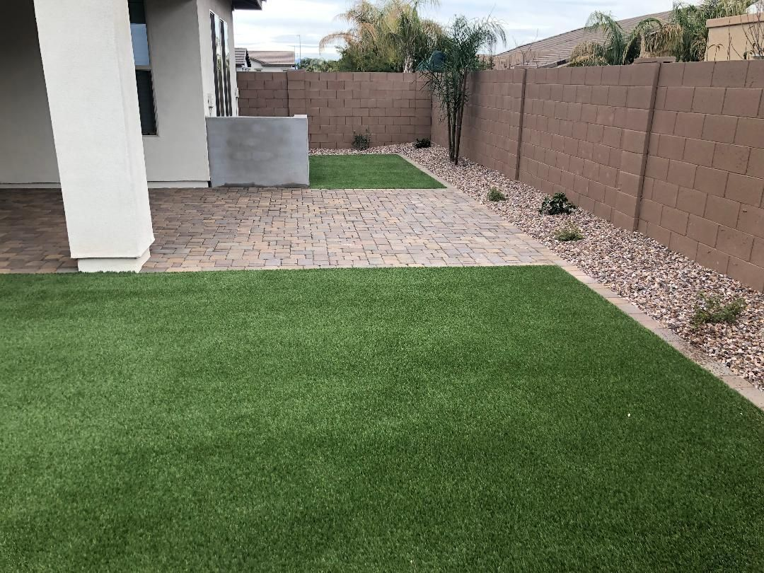 Backyard with green turf, brick patio, and brown walls. Small plants in rock bed.