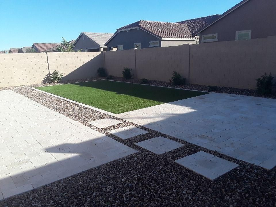 Backyard with stone patio, gravel border, grass, and tan walls under a blue sky.