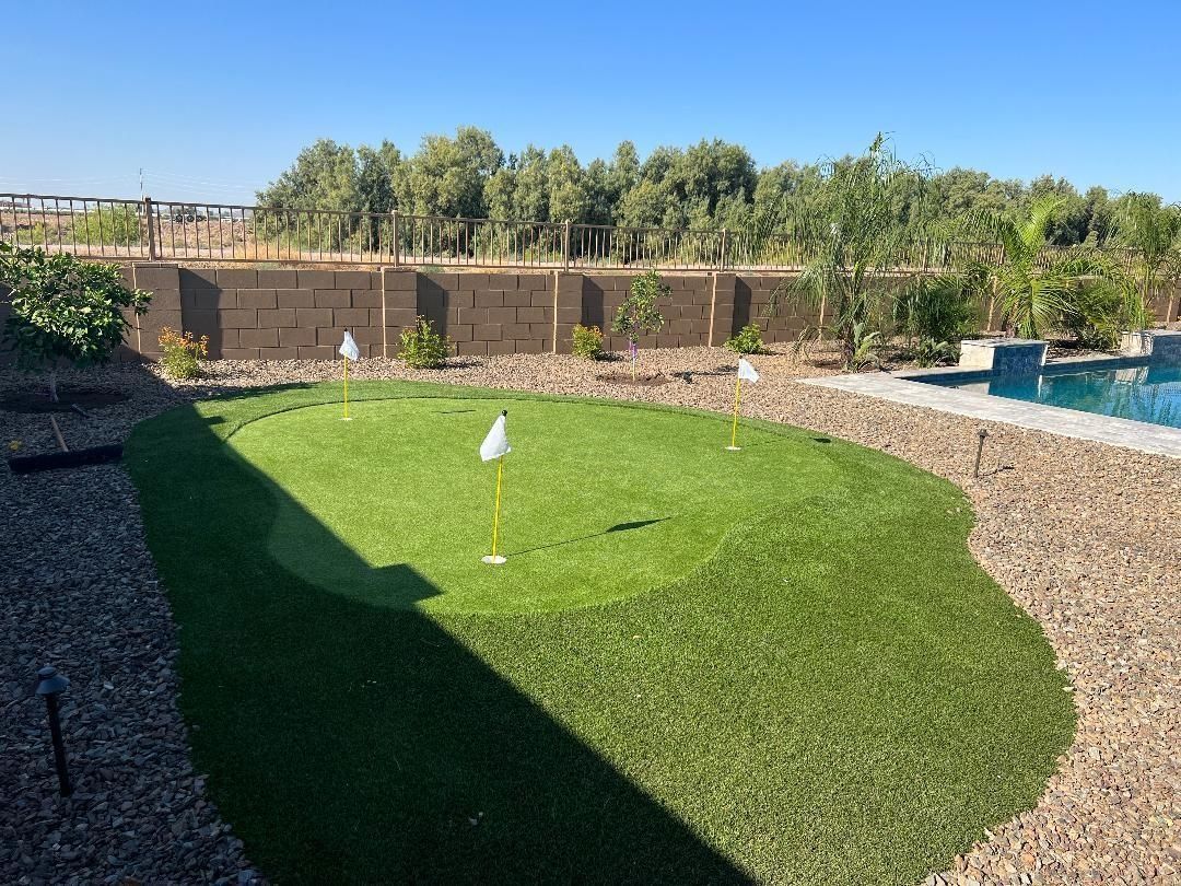 A backyard putting green with a flagstick, bordered by rocks and a pool on a sunny day.