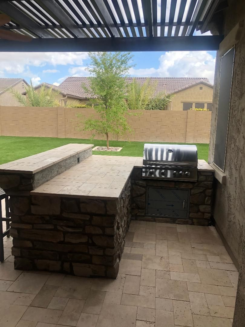 Outdoor kitchen with built-in grill and counter, stone facade, under a pergola, overlooking a backyard with grass and fence.