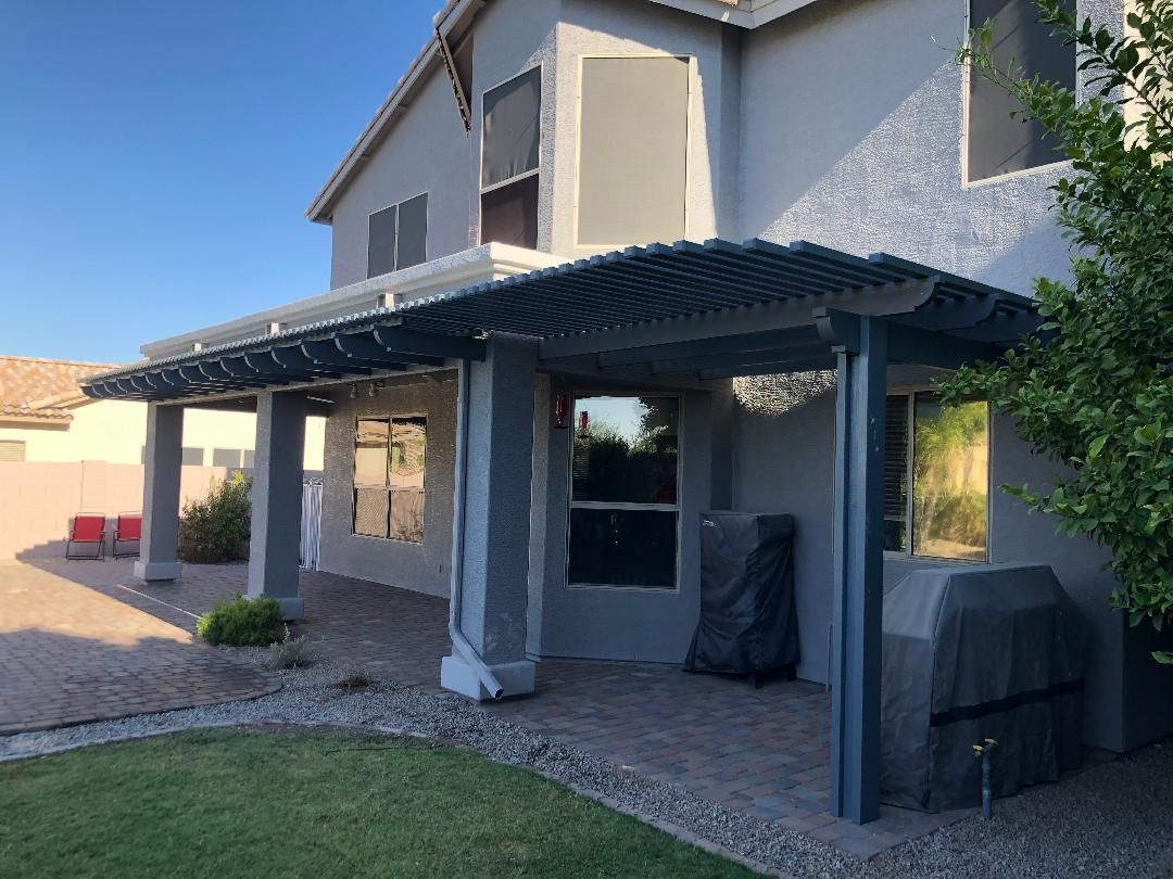 Gray pergola attached to a two-story beige house with window shades. A grill is covered nearby.