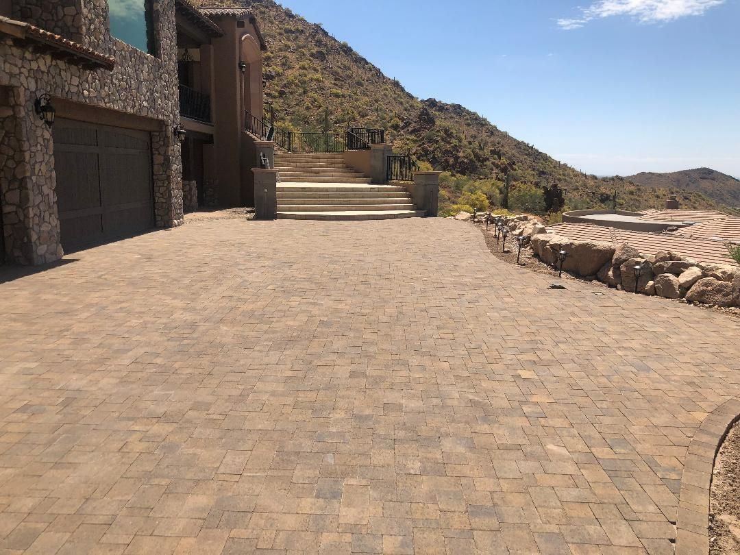Stone-paved driveway leading up to a house with stone siding against a hillside. Sunny, clear sky.