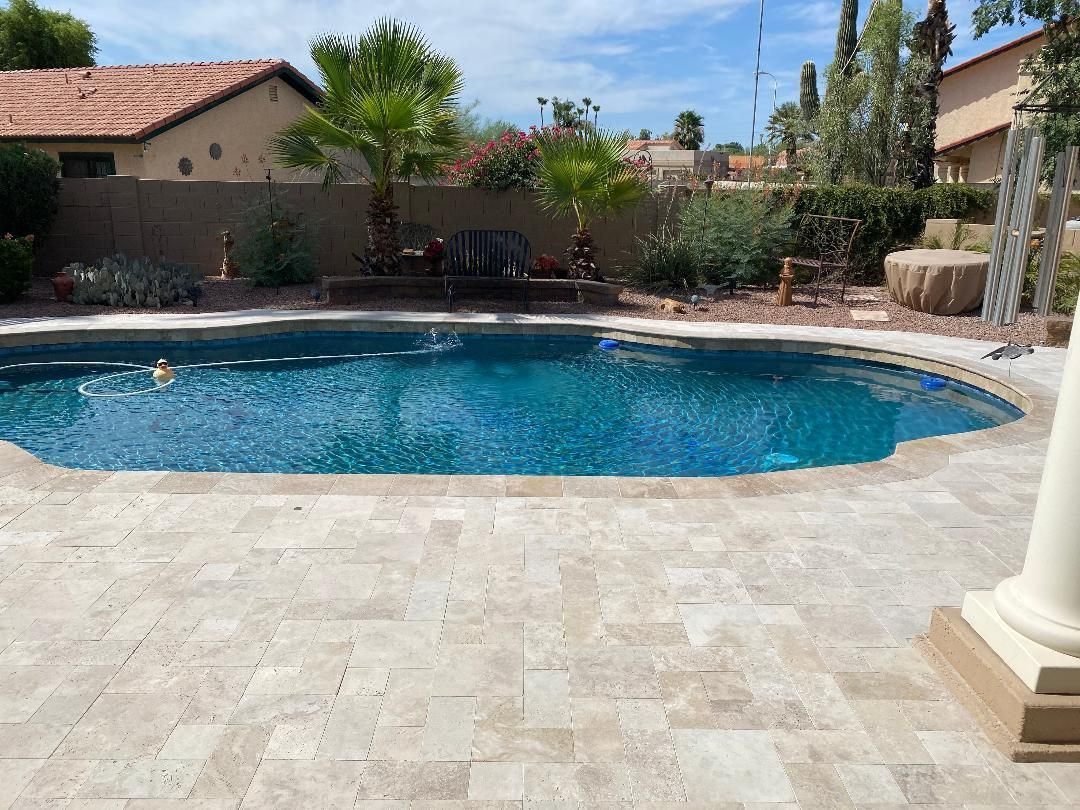 Swimming pool with blue water surrounded by beige stone patio, with palm trees and a house in the background.