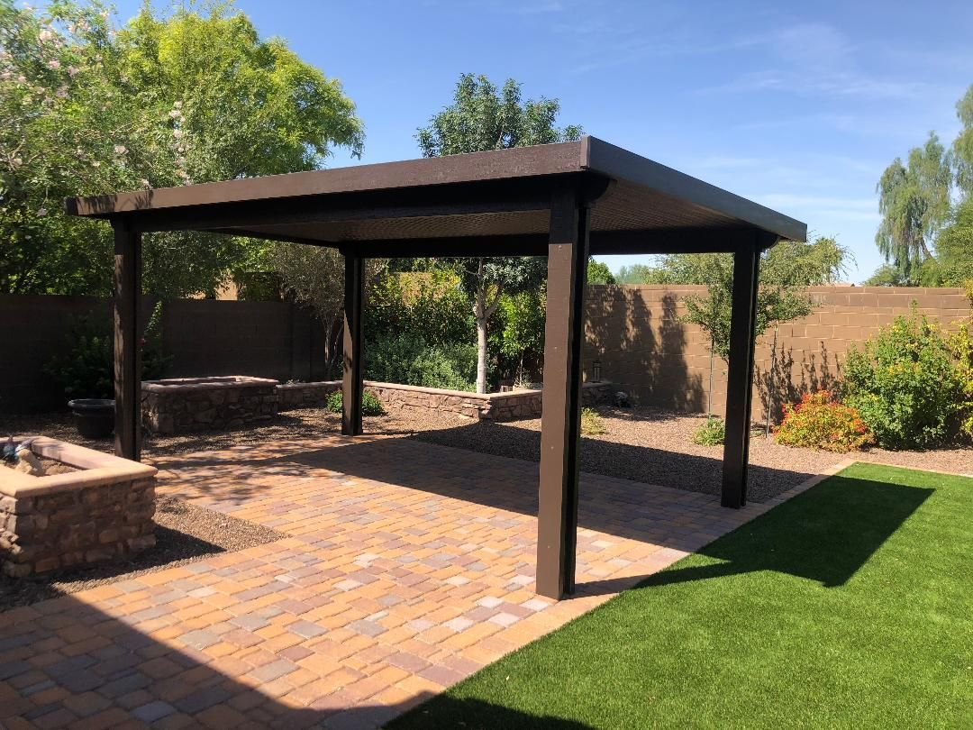 Brown pergola on a brick patio with green grass and trees in the background. Sunny day.