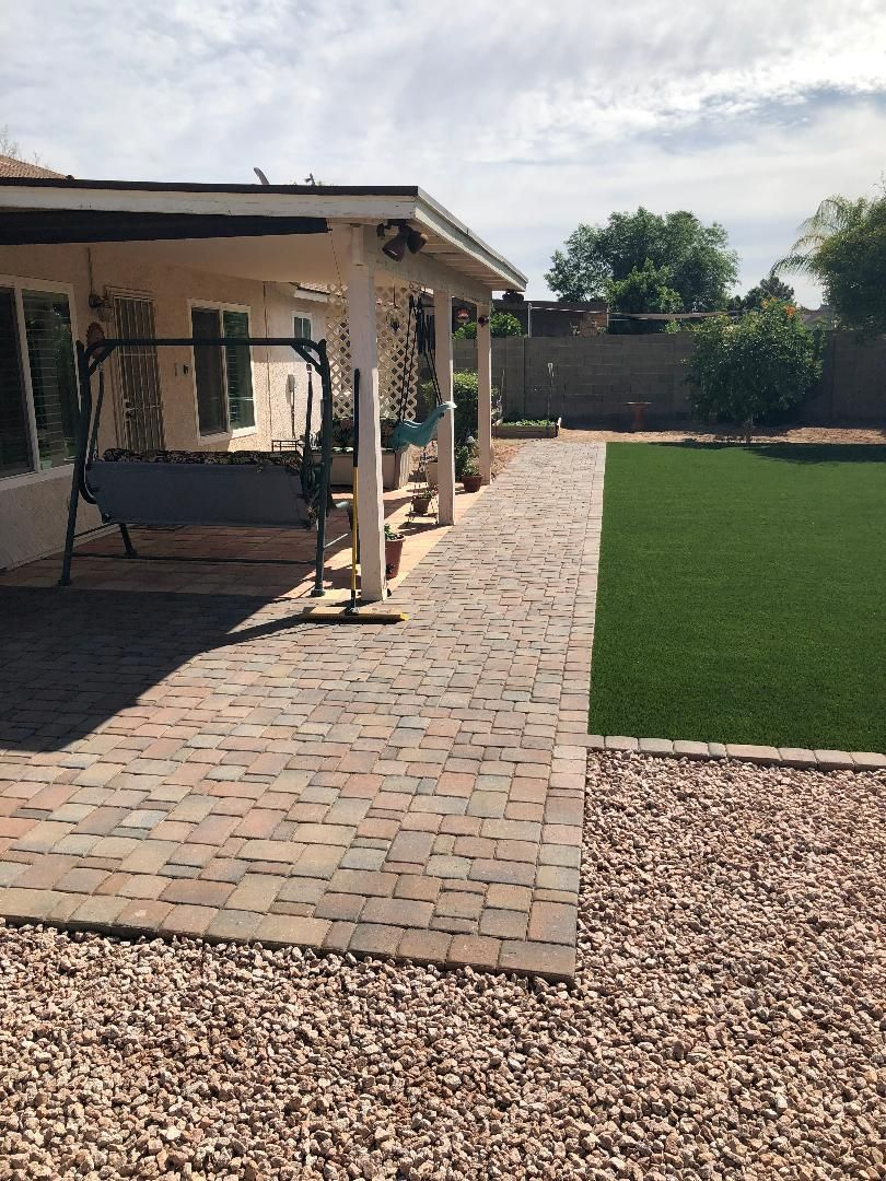 Brick pathway leading past a porch with swing and into a green lawn, gravel border, sunny outdoor setting.