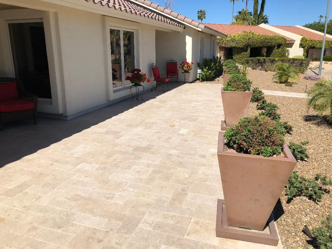 A paved patio next to a cream-colored building. Brown planters with green plants in the foreground, red chairs in the background.