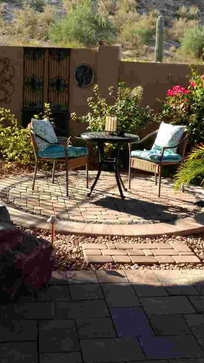Patio with two chairs, small table, and circular brick area against a wall with plants, under a sunny sky.
