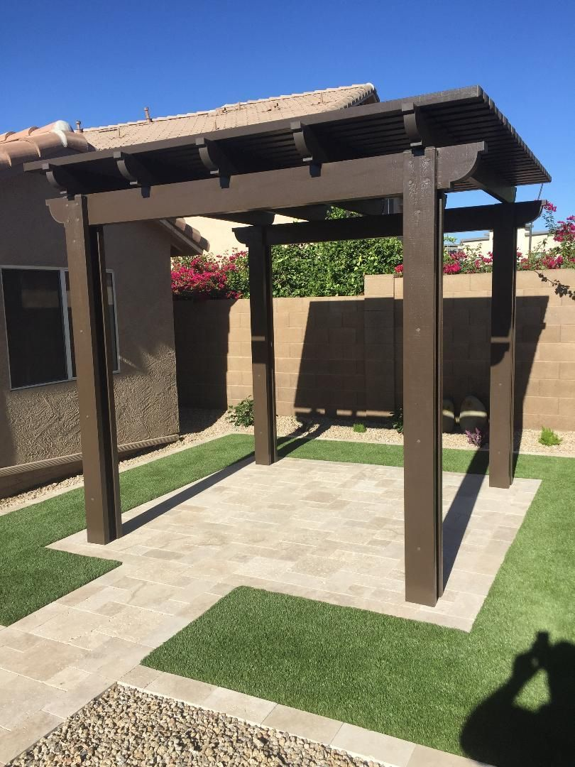 Brown wooden pergola on a stone patio with surrounding green grass and a house in the background.