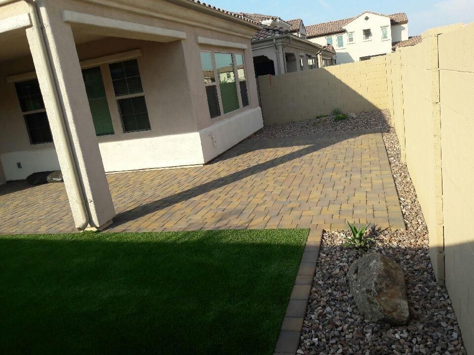 Backyard patio with brick pavers, lawn, and rock border alongside a tan wall.
