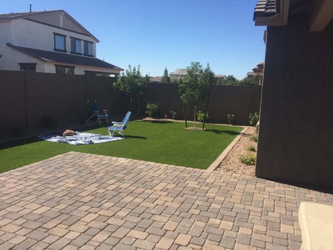 Backyard with a brick patio, green lawn, and brown fence under a blue sky.