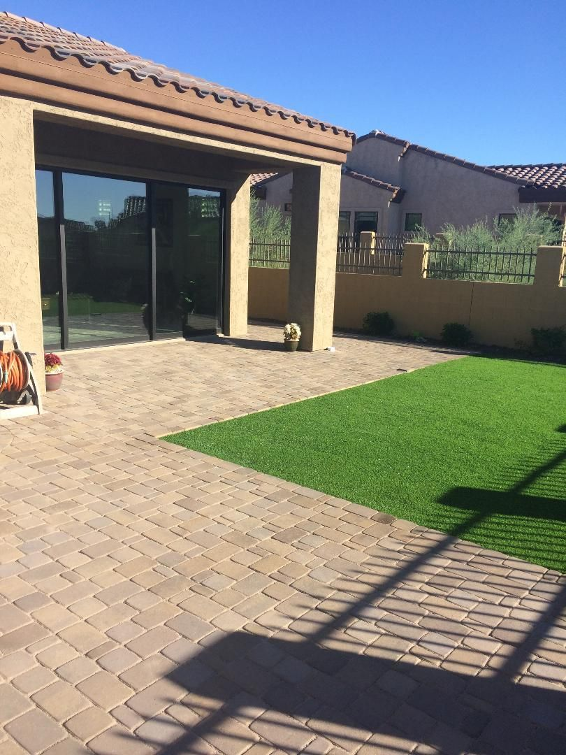 Backyard patio with brick pavers, green turf, and partial shade. A house is in the background.