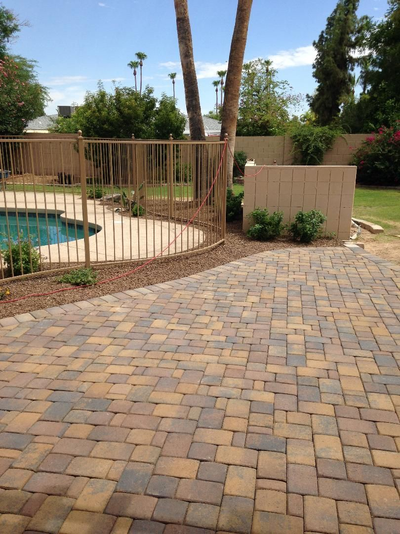 Brick patio leading to a fenced pool area with trees and a light-colored wall in the background.