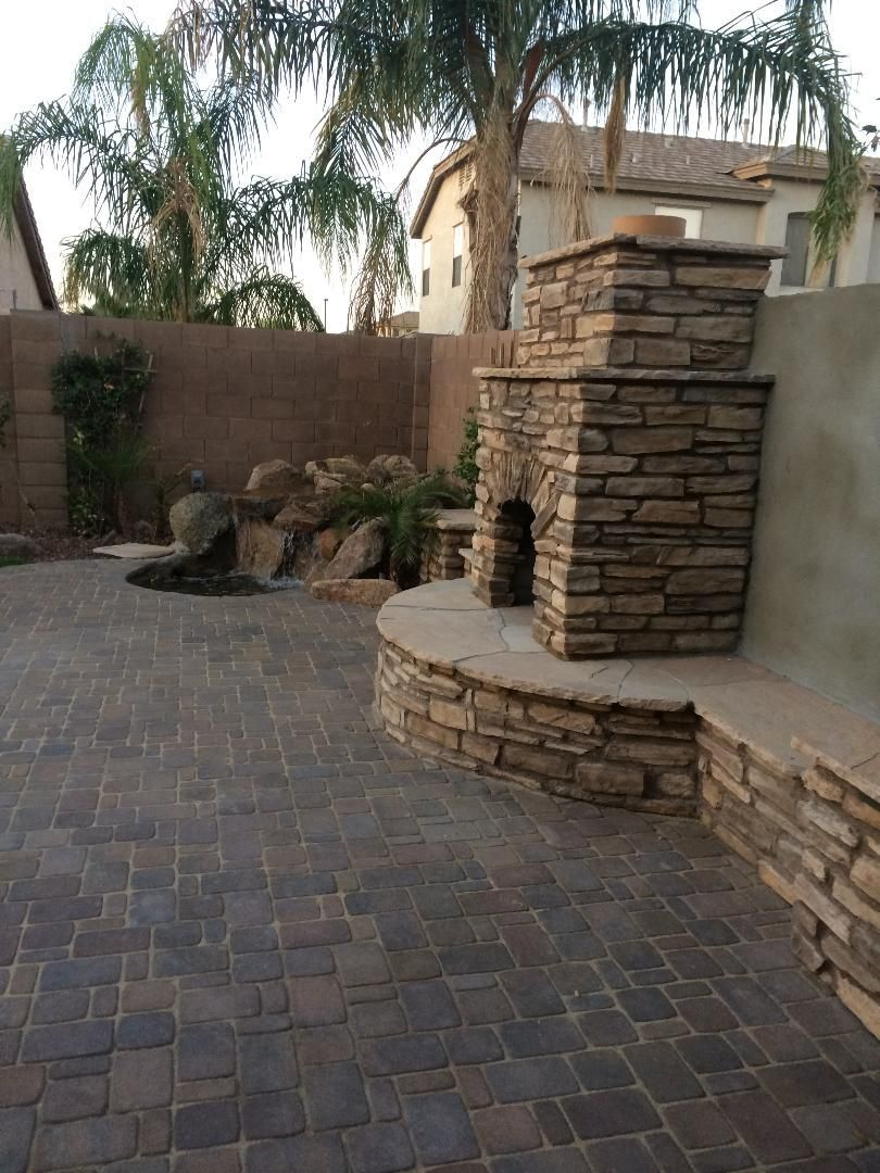 Patio with stone fireplace, waterfall, and brick paving, in a backyard setting.