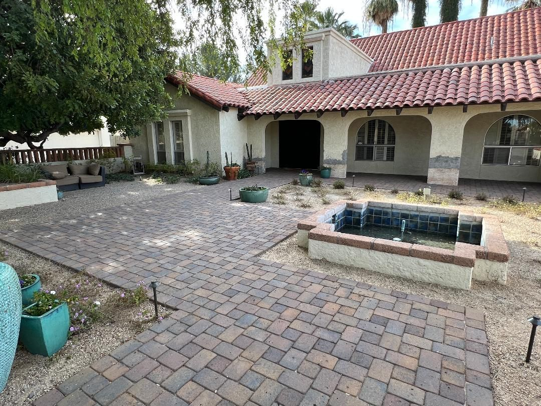 Brick-paved patio in front of a Spanish-style house with terracotta roof. Includes a water feature and sparse landscaping.