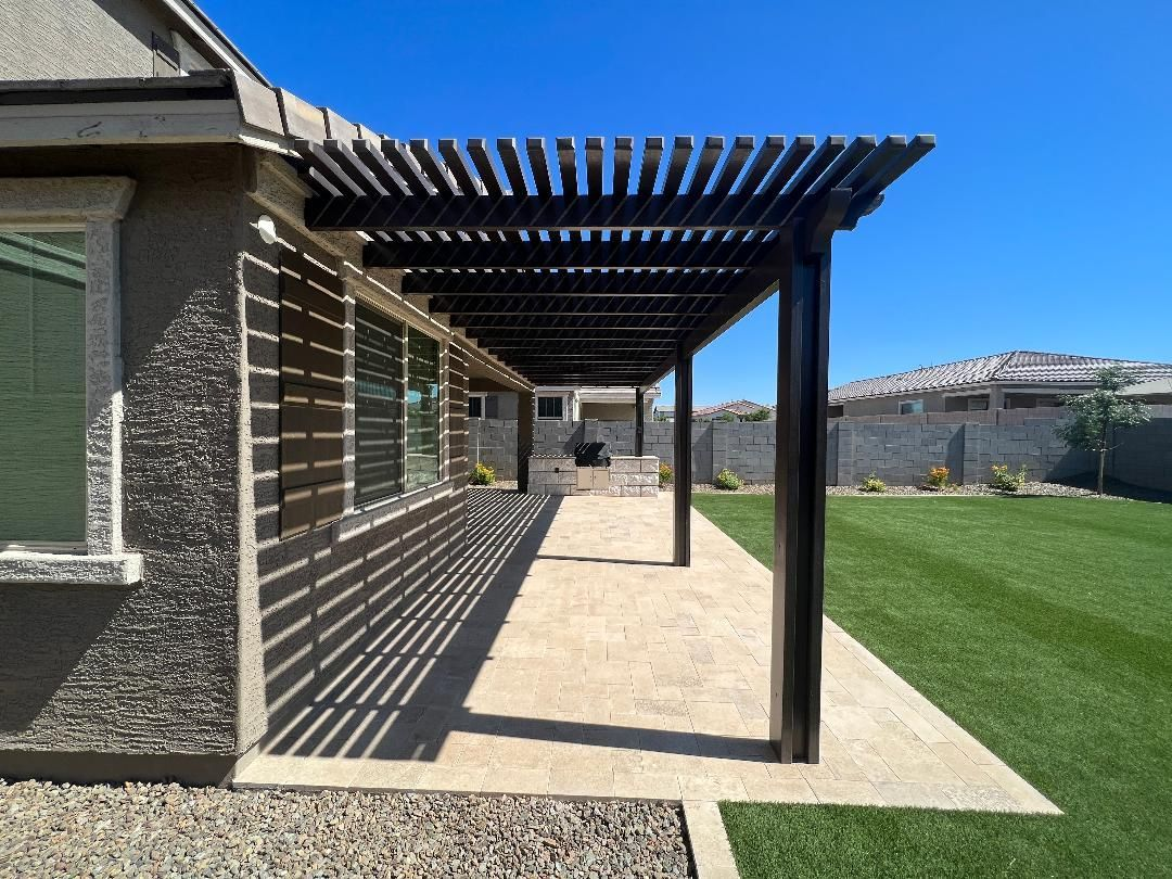 Patio with dark pergola, casting shadows on the concrete floor. Green lawn and clear blue sky.