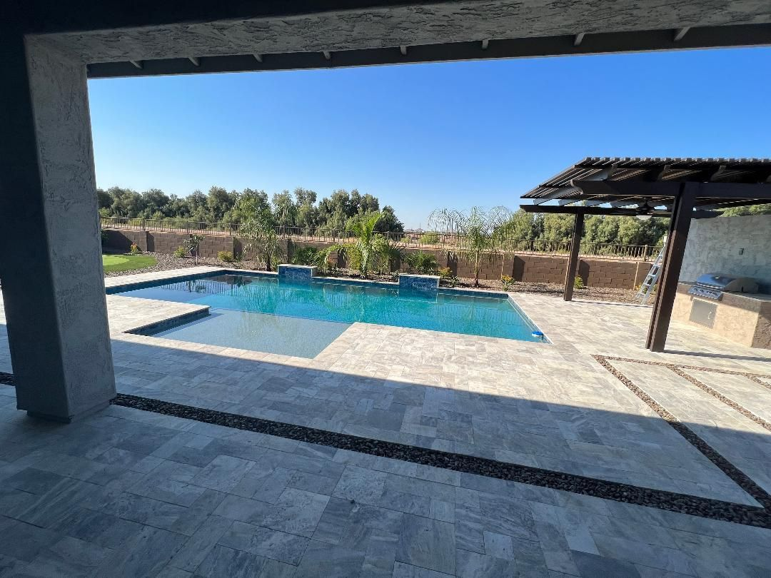 View of a pool and outdoor space framed by a patio. The pool is blue and sits next to a shaded area.