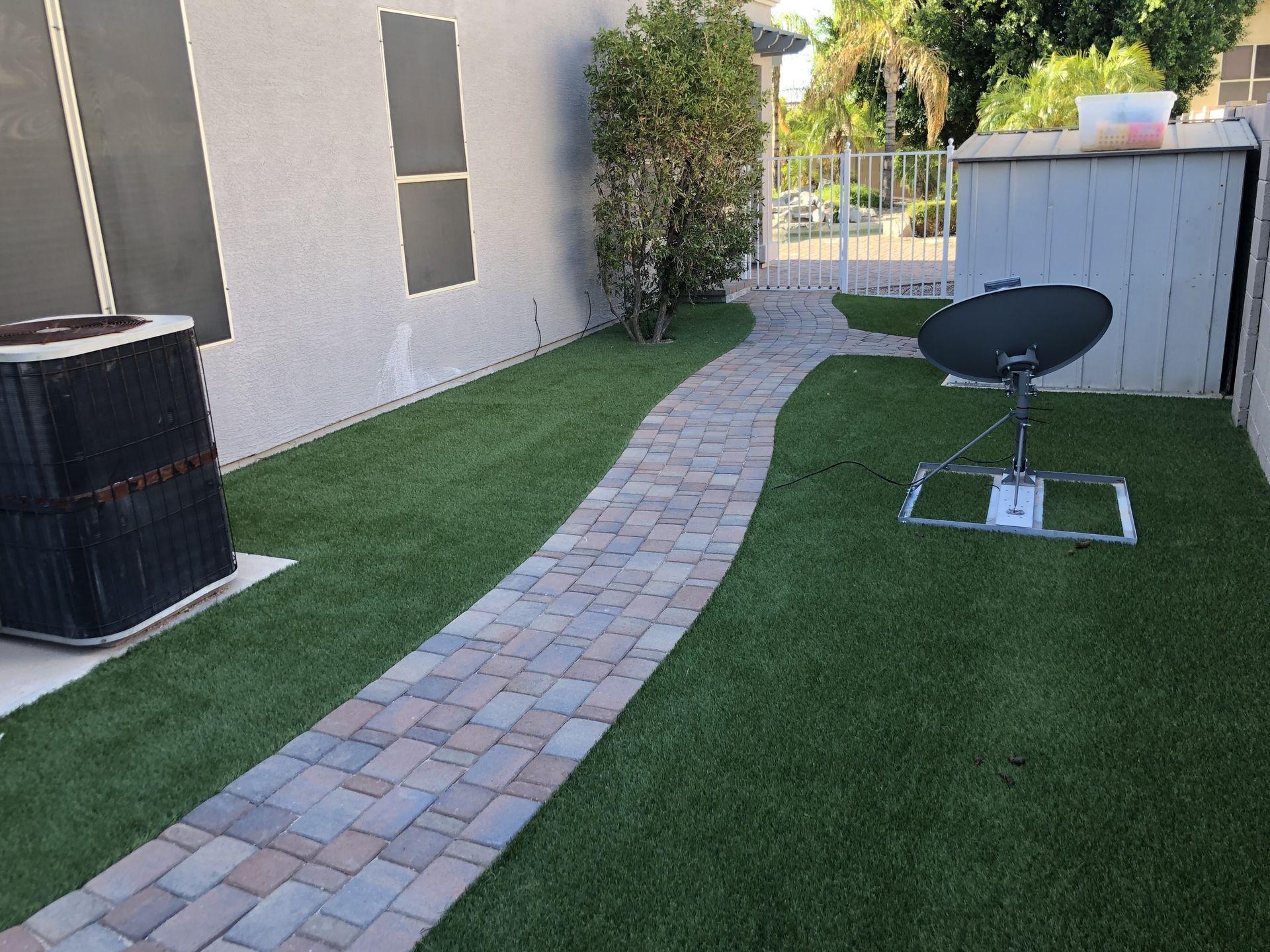 A brick pathway winds through a grassy yard with an AC unit, satellite dish, and shed.