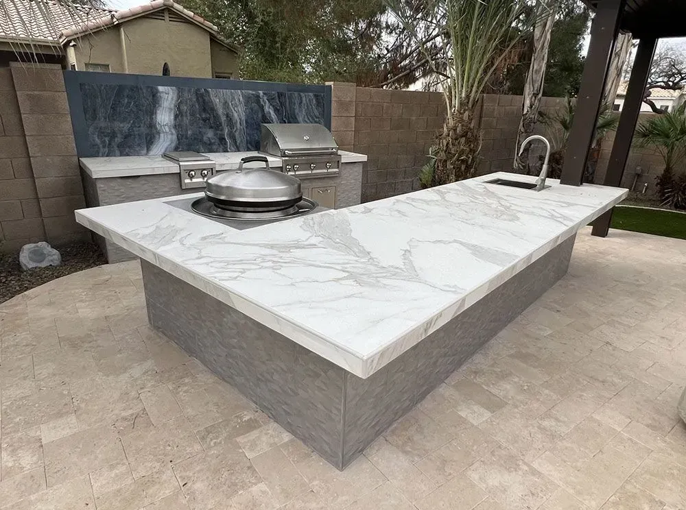 Outdoor kitchen with marble countertops, stainless steel grill, and sink. Grey tiled base, beige patio.