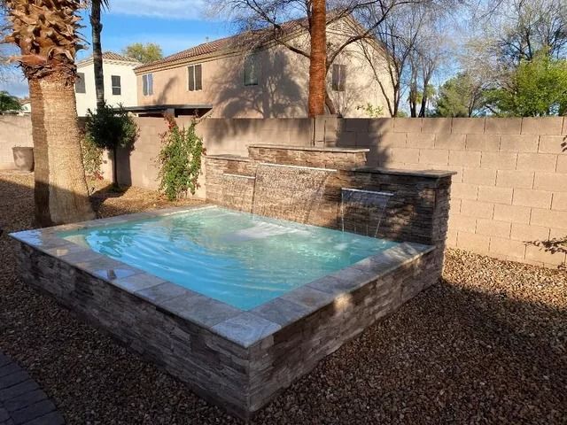 Rectangular hot tub with waterfall feature in a backyard with stone wall and gravel.