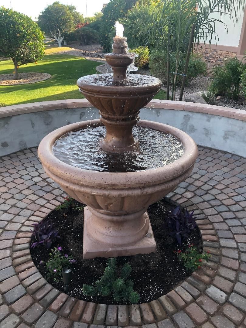 Fountain with tiered basins, water spraying upward. Surrounded by brick paving and small garden, outdoors.
