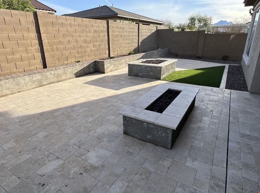 Backyard patio with two rectangular fire pits on light stone pavers, surrounded by a beige wall.