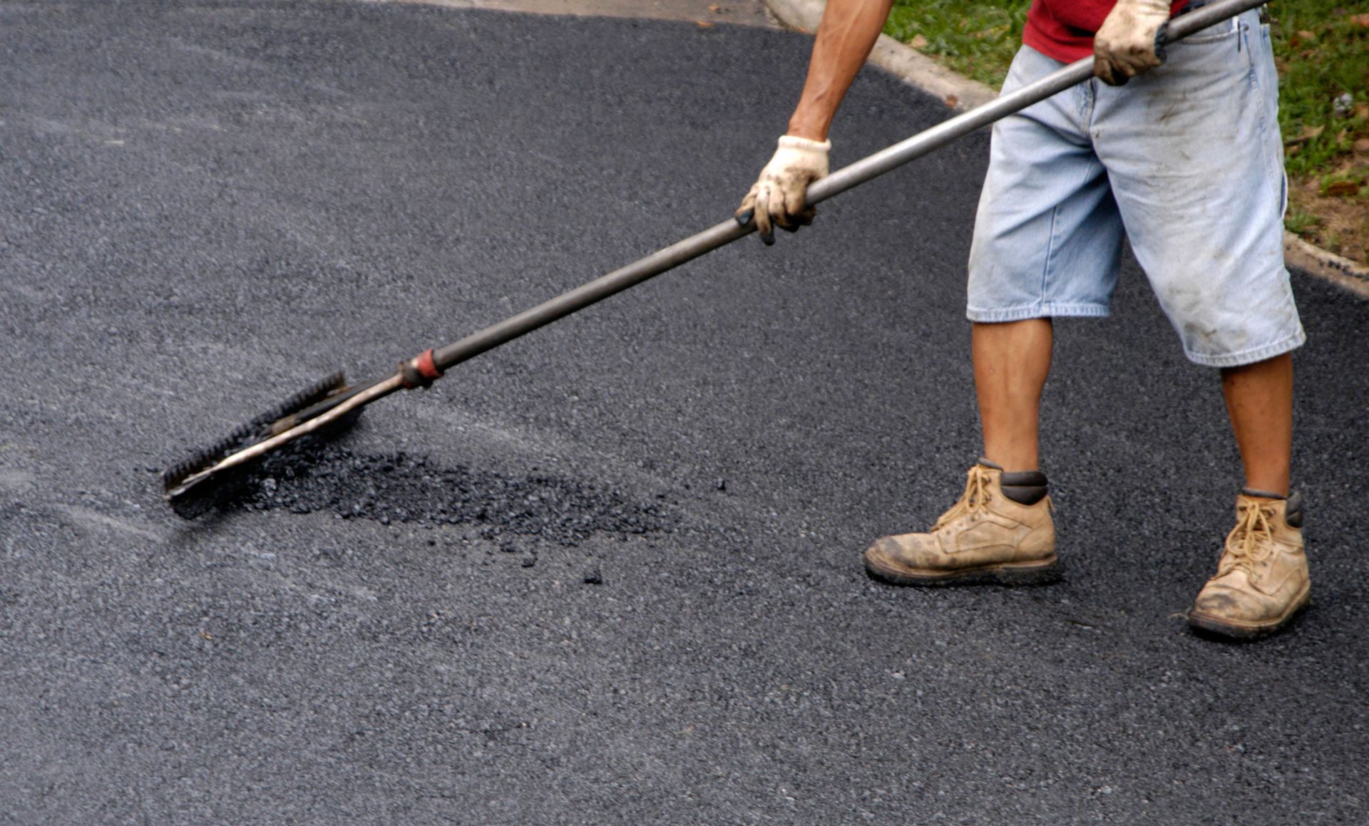 Person smoothing fresh asphalt with a long-handled tool, wearing gloves, work boots, and shorts.