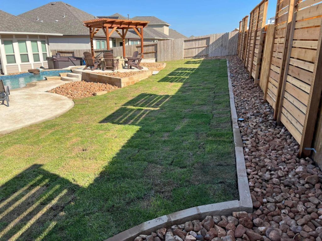 Backyard with new grass, a patio with a pergola, a pool, and a rock bed next to a wooden fence.