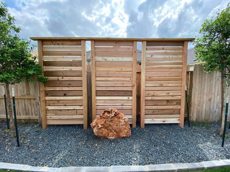 Wooden privacy screen with a large boulder in a gravel bed. Trees on either side.
