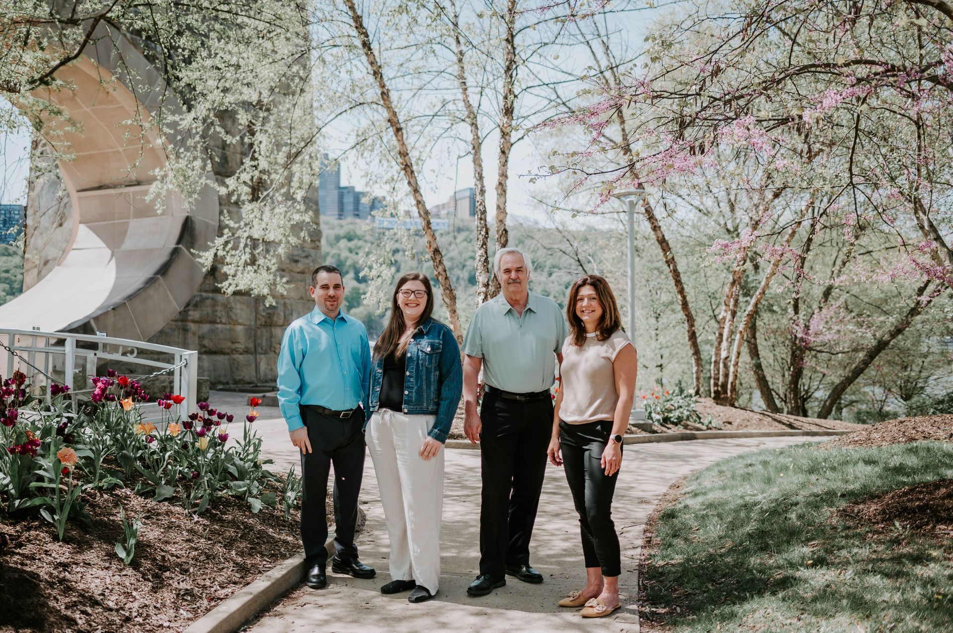 Four people posing outdoors in front of a stone structure and flowering trees. They are smiling, dressed in casual clothing.