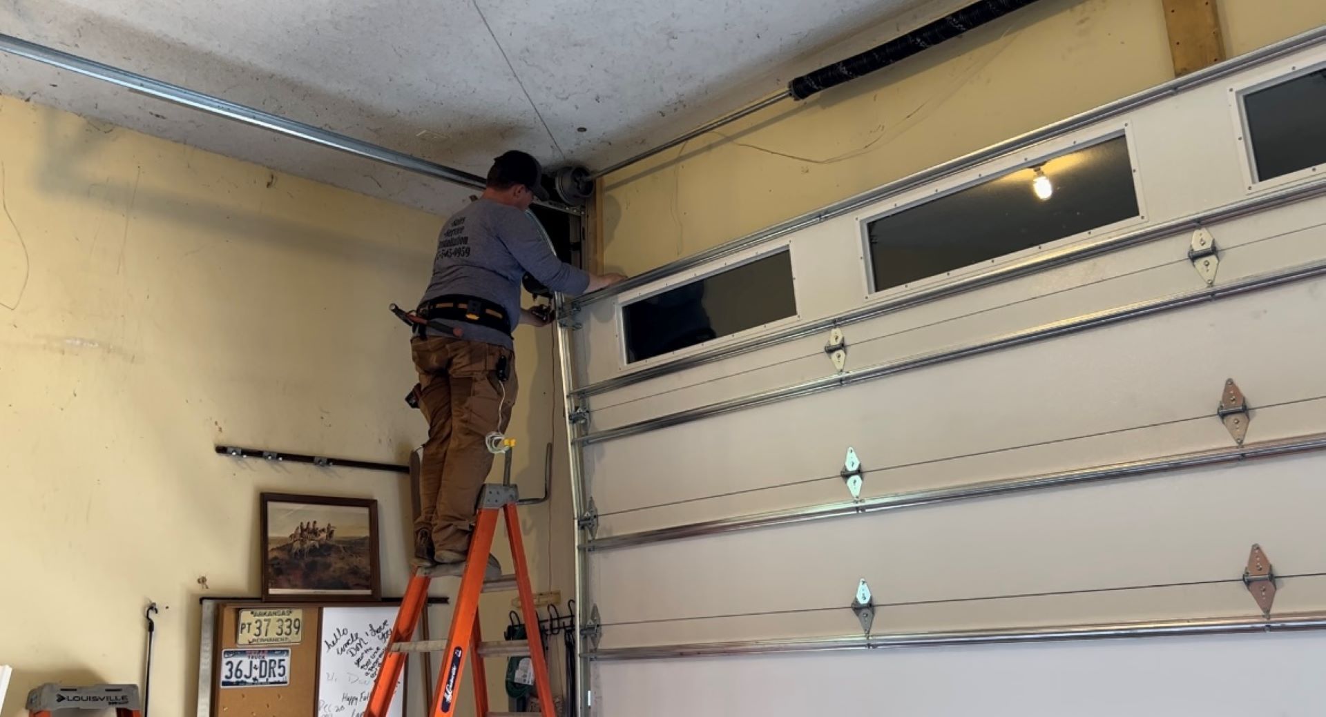 A person on a ladder repairs a garage door in a garage.