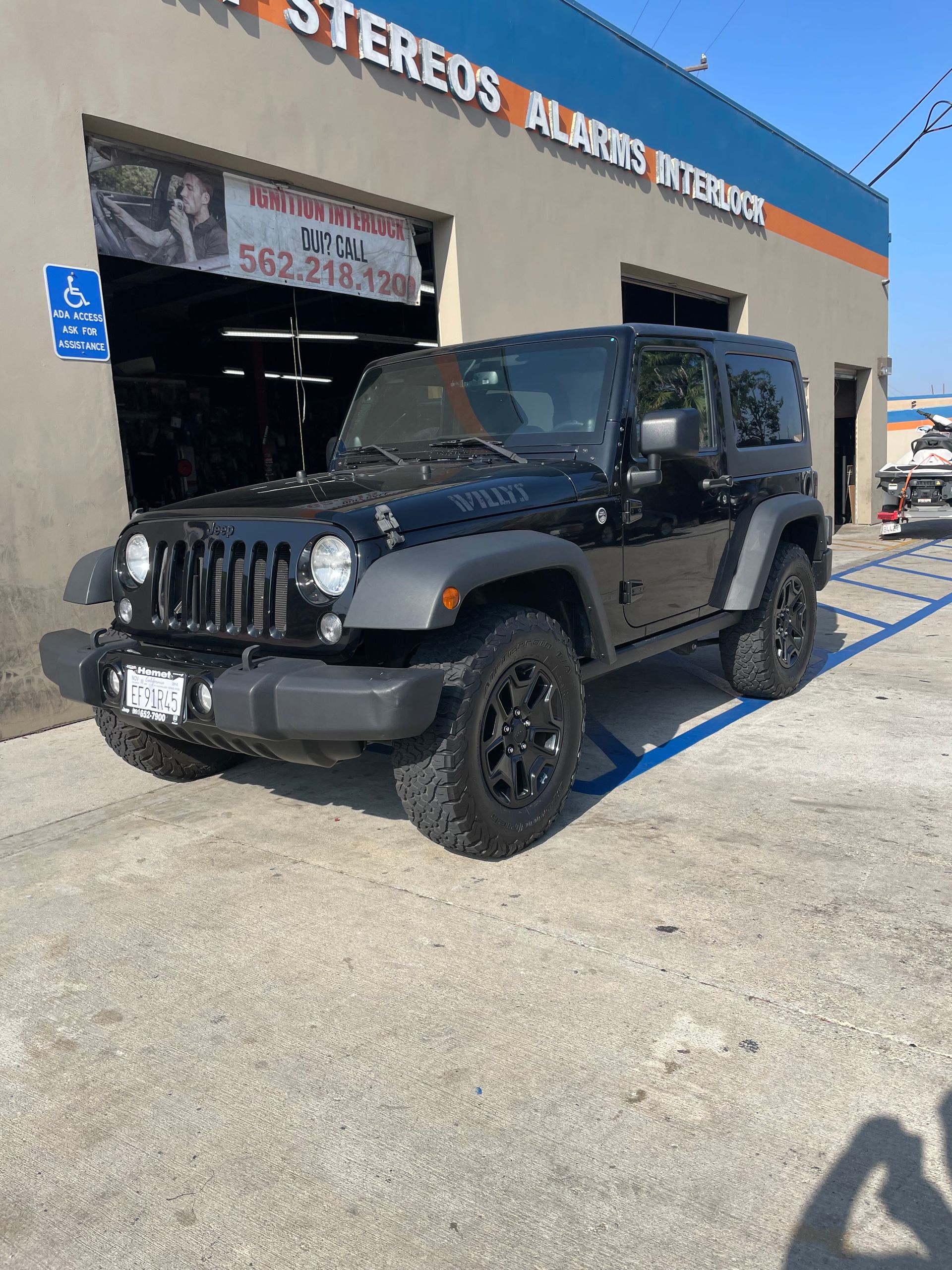 Black Jeep Wrangler parked in front of a building with a blue awning, on a sunny day.