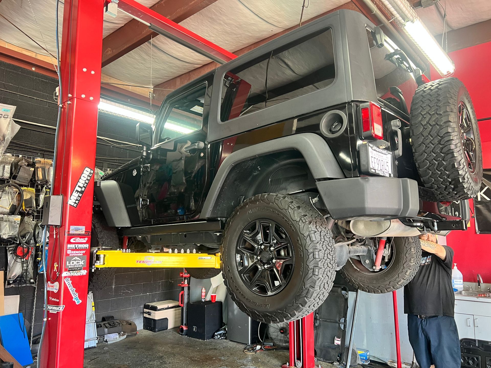 Black Jeep Wrangler on a lift in a repair shop. A person stands nearby.