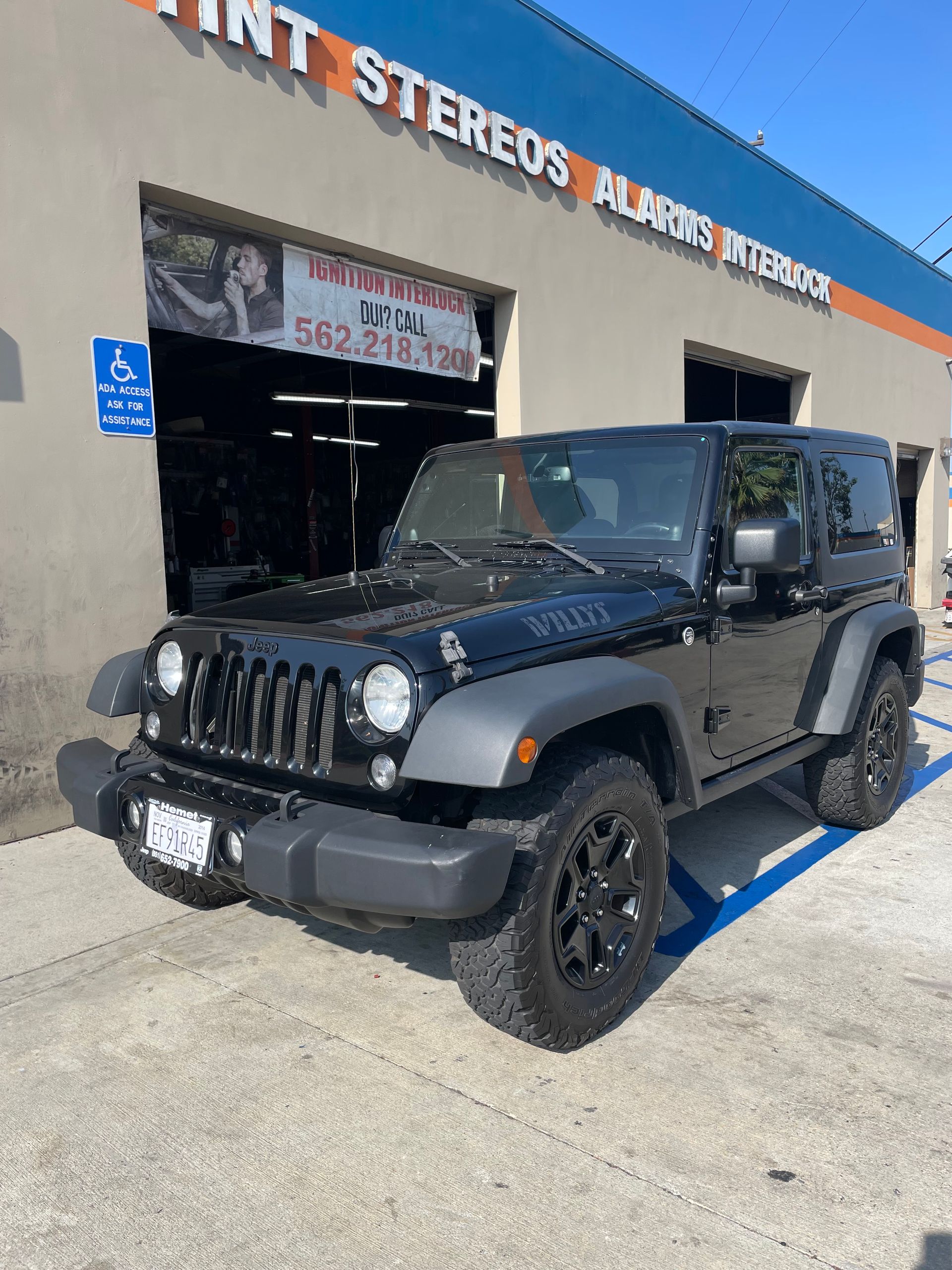 Black Jeep parked in front of a business with tinted windows, stereos, and alarms. Blue handicap sign.