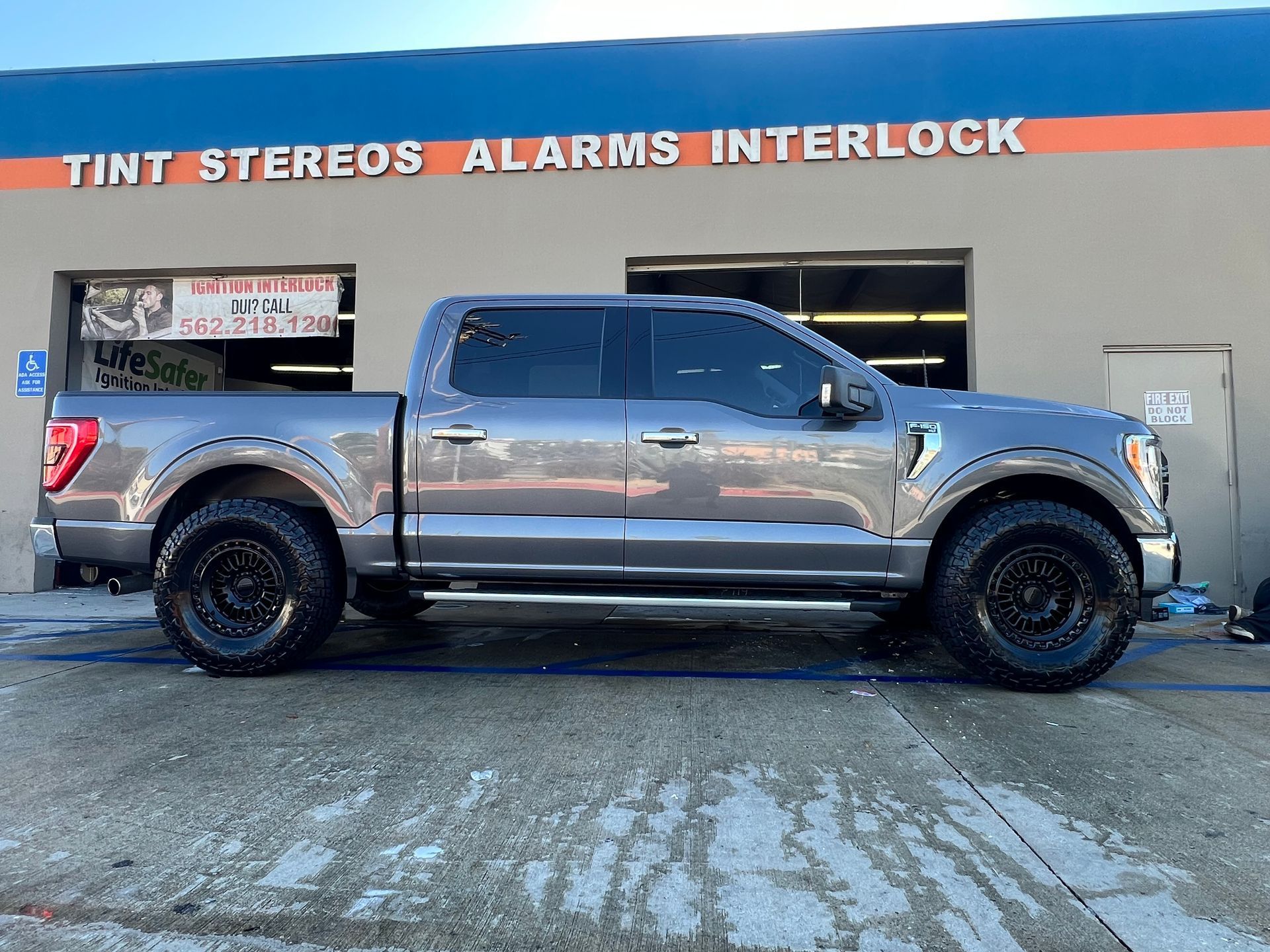 Gray pickup truck with tinted windows parked in front of a shop advertising tint, stereos, alarms, and interlock services.