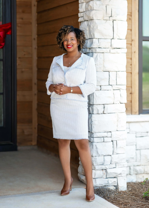 Woman in white suit stands by a stone pillar, smiling.