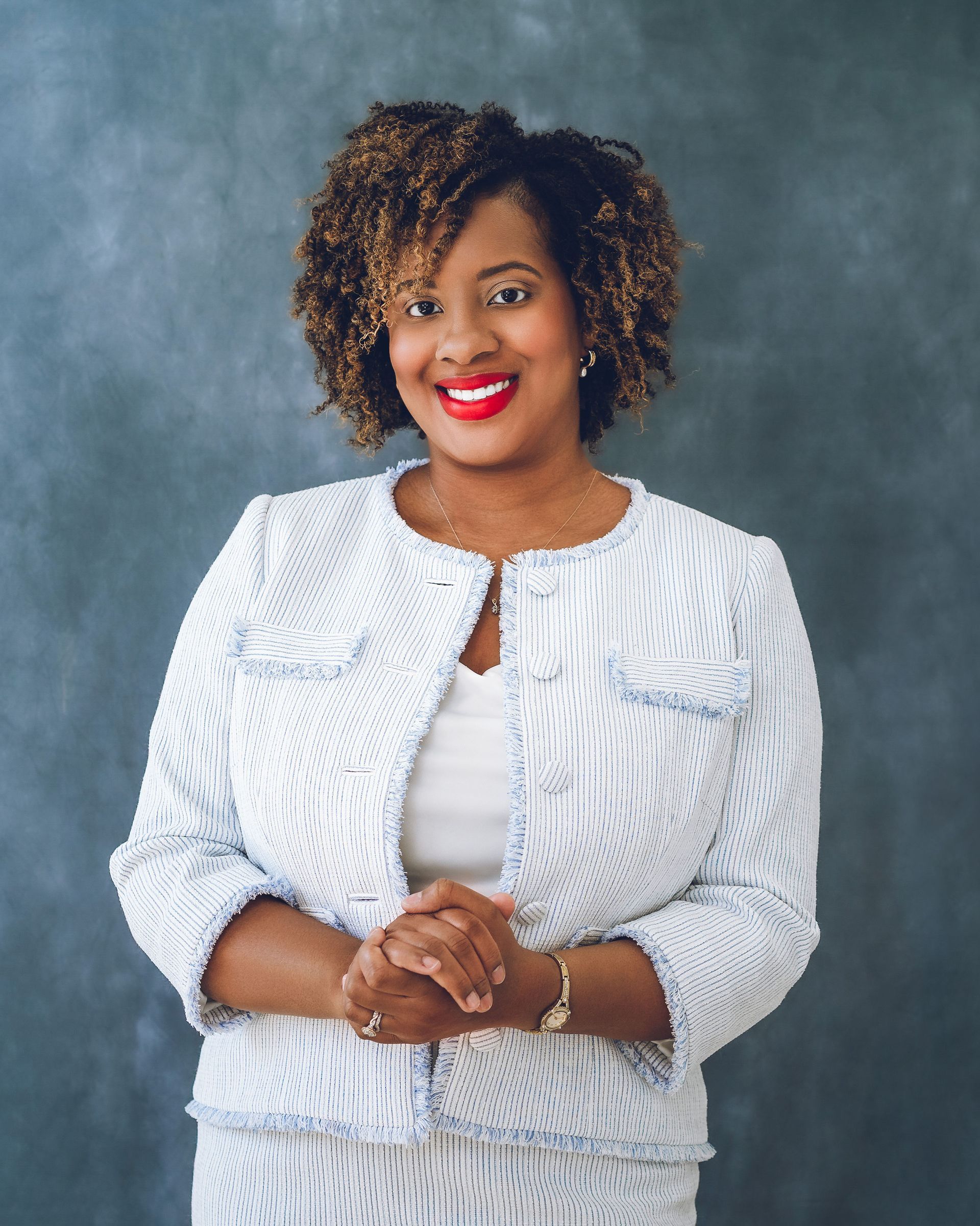 Woman in white patterned suit, hands clasped, smiling with red lipstick, against a gray background.