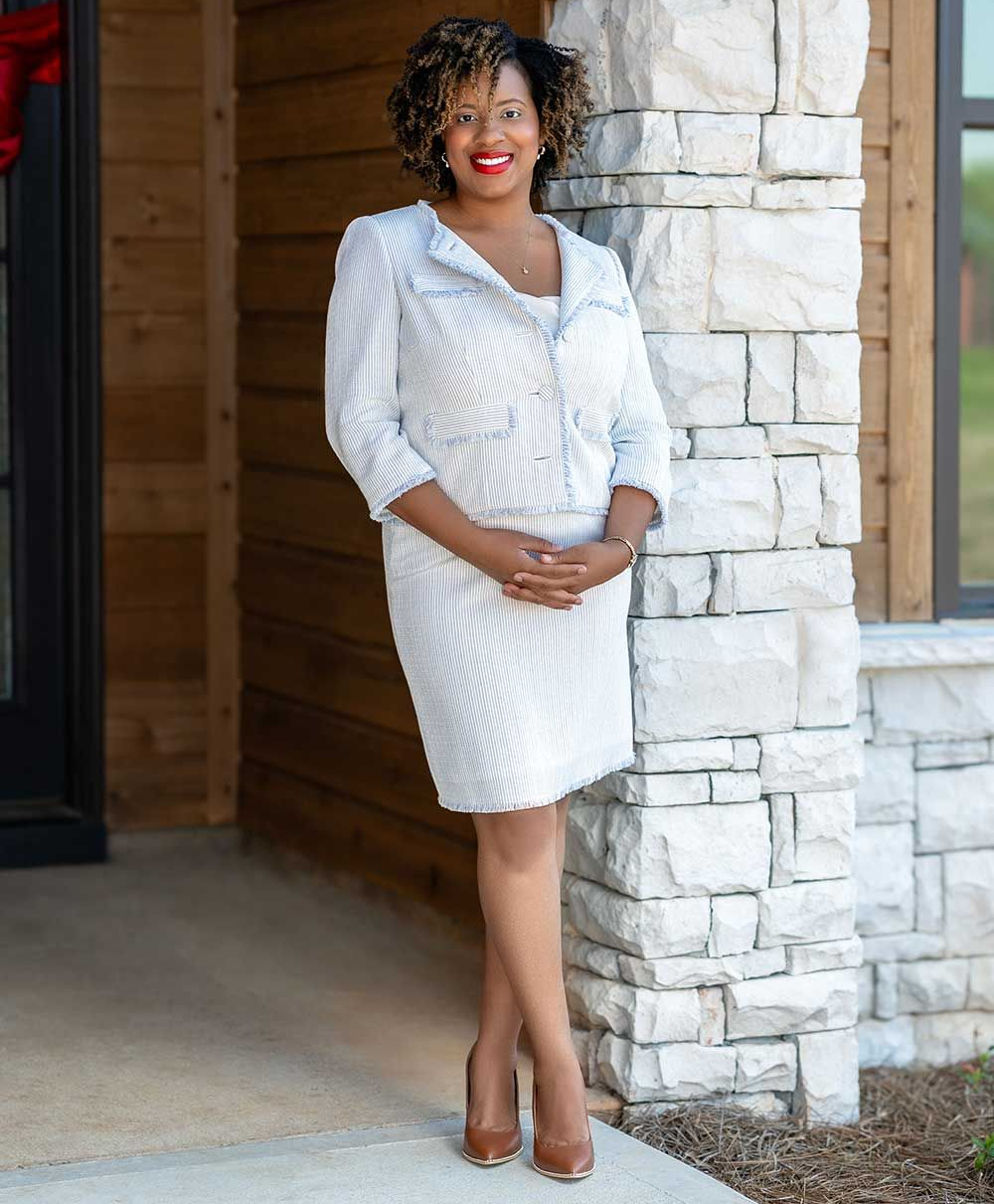 Woman in white suit stands by stone column, smiling, outdoors.