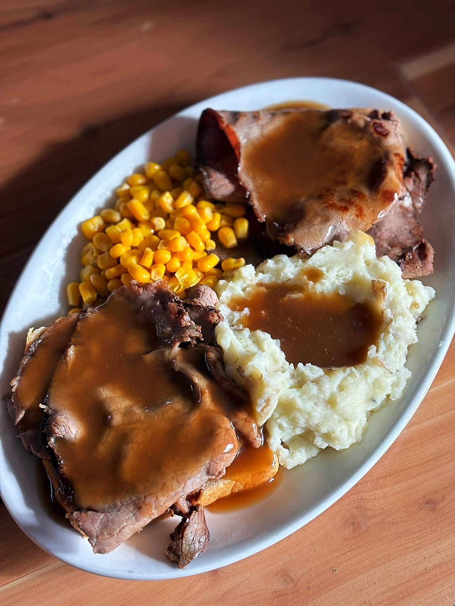A plate of roast beef , mashed potatoes , corn and gravy on a wooden table.