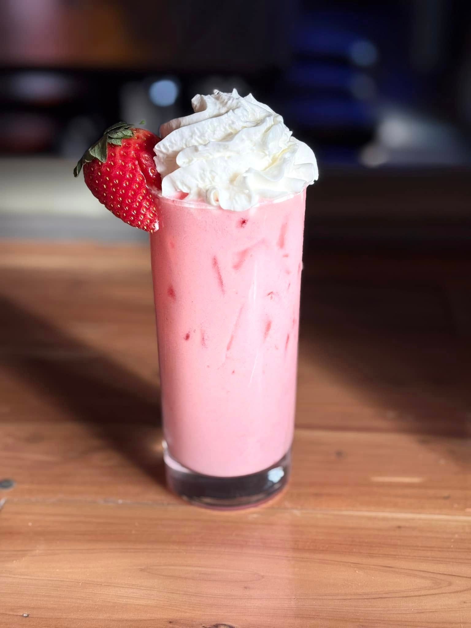 A pink milkshake with whipped cream and a strawberry on top on a wooden table.