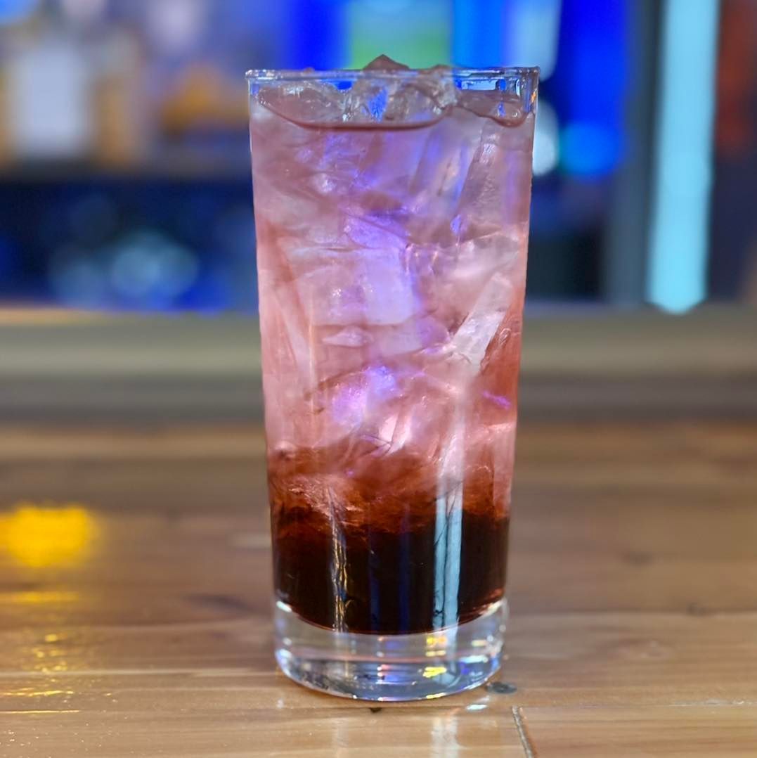 A tall glass filled with pink liquid and ice is sitting on a wooden table.