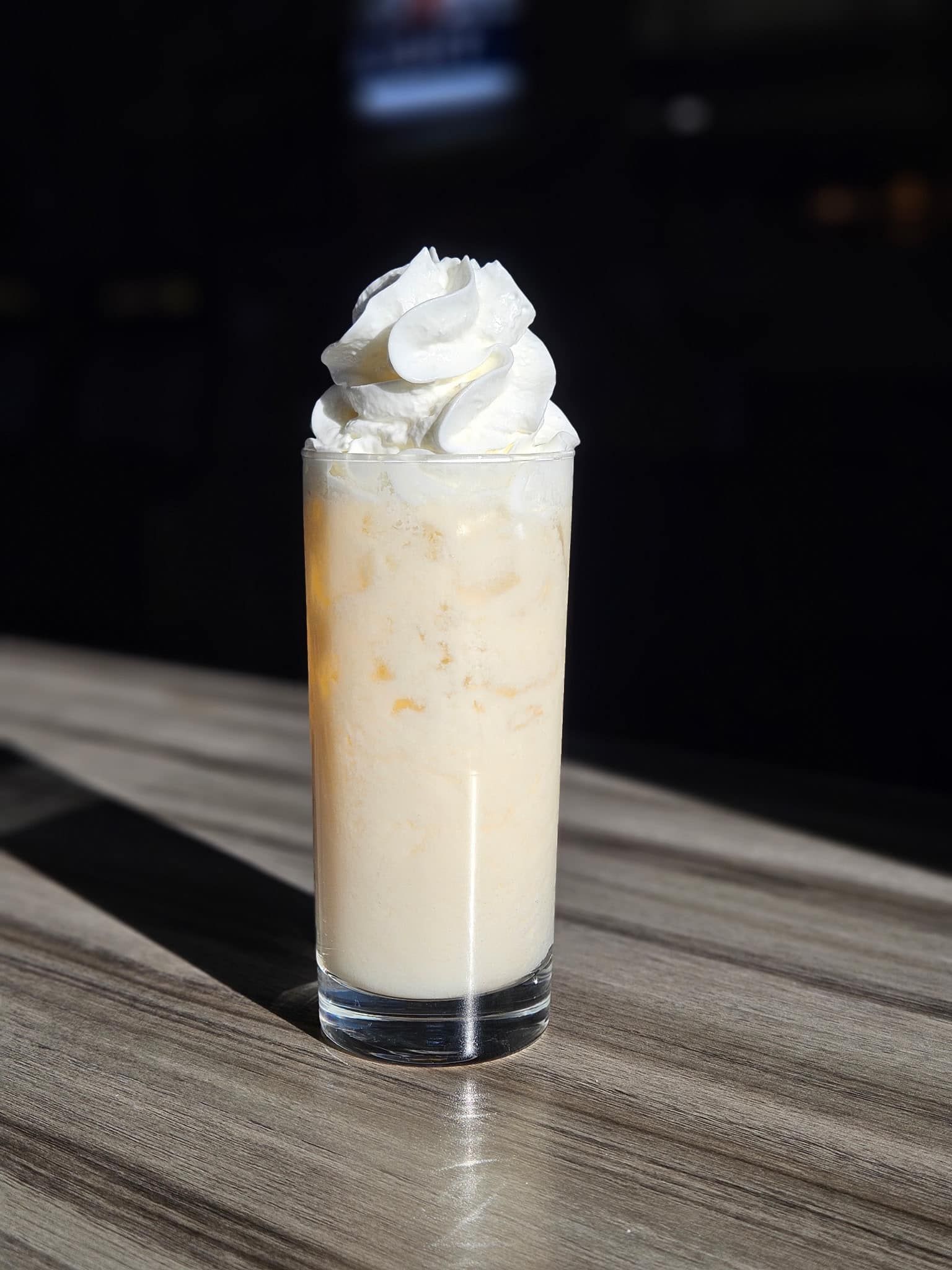 A glass of milkshake with whipped cream on top is on a wooden table.