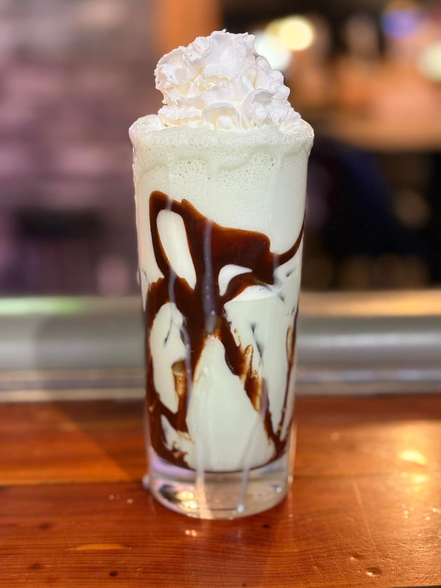A milkshake with whipped cream and chocolate sauce is on a wooden table.
