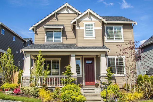Two-story beige house with red front door, white trim, and a small front porch surrounded by greenery